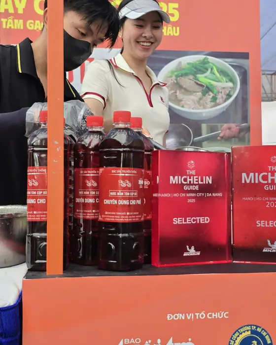Chai Tôm Cọp Đỏ cạnh biển Michelin Guide Selected 2025, phía sau là nhân viên gian hàng và băng rôn báo Tuổi Trẻ