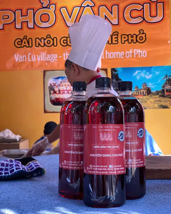 Ba chai Tôm Cọp Đỏ trên bàn trước băng rôn Phở Vân Cù, cái nôi của nghề phở, phía sau là đầu bếp đội mũ trắng
